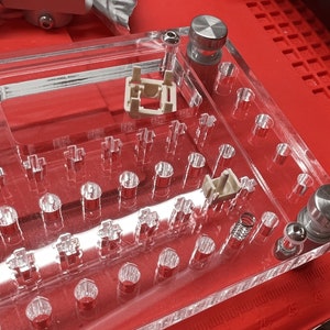 May include: A clear acrylic keyboard base with silver-colored metal components and key switch housings. The base is on a red surface, with various holes for key switches and stabilizers. The design is for a custom mechanical keyboard.