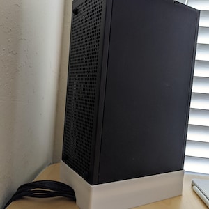 May include: A black computer case with a perforated front panel and a solid side panel. The case sits on a white base and has a black power cord. The case is on a light wood surface.