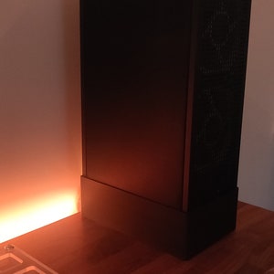 May include: A black computer case with a mesh side panel sits on a wooden surface. The case is rectangular and has a minimalist design. A white stylus and a clear acrylic component are also on the table.