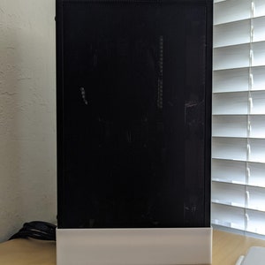 May include: A black computer case with a mesh front panel and a white base. The case is rectangular and stands upright on a light-colored wooden surface. The background includes a window with white blinds.