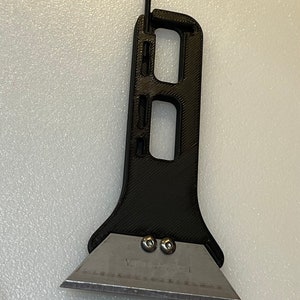 May include: A black utility knife with a metal blade and a black handle. The handle has a unique design with a built-in hex key. The blade is secured with two silver screws. The tool is designed for cutting and scraping.