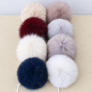May include: A collection of eight fluffy pom-poms arranged in two columns. The colors include cream, dusty rose, deep burgundy, light gray, off-white, mauve, navy blue, and bright white. Each round pom-pom features soft, dense fibers.