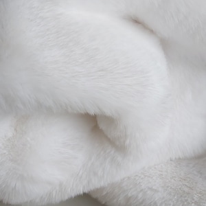 Może przedstawiać: Zbliżenie miękkiej, białej, puszystej tkaniny. Materiał ma gęstą, pluszową fakturę z długimi, płynącymi włóknami. Tkanina jest złożona, tworząc cienie i głębię, podkreślając jej luksusowy wygląd.