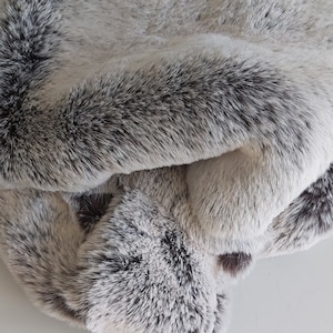 Pode incluir: Um cobertor de pele sintética dobrado em tons de cinza e branco. O material de pelúcia tem um padrão salpicado com detalhes em cinza escuro, criando uma aparência macia e texturizada. O cobertor está dobrado sobre si mesmo, mostrando a textura fofa.