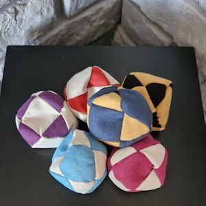 May include: Six colourful fabric juggling balls. The balls are made of different colours of fabric, including red, white, blue, yellow, purple, and black. The balls are arranged in a circle on a black surface.