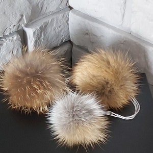 May include: Three fluffy pom poms in different shades of brown. The pom poms are made of fur and are attached to strings.