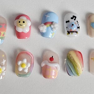 Könnte beinhalten: Ein Set aus zehn künstlichen Nägeln mit verschiedenen Designs. Die Nägel weisen farbenfrohe Muster auf, darunter Kirschen, Blumen, Regenbogen und Zeichentrickfiguren. Jeder Nagel hat ein einzigartiges Design mit glänzendem Finish.