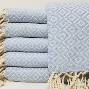 May include: A stack of light blue and white diamond-patterned Turkish towels with cream-colored fringe. The towels are folded and stacked, showcasing the geometric design. The soft color palette and texture suggest a spa or beach theme.