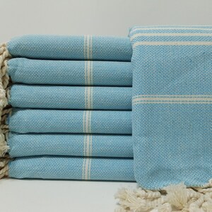 May include: A stack of light blue Turkish towels with white stripes and fringe. The towels are folded and stacked on a white surface.