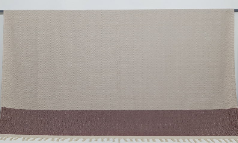 K&ouml;nnte beinhalten: Eine gewebte Wolldecke in Beige und Braun mit Fransenrand. Die Decke hat ein dezentes Rautenmuster.