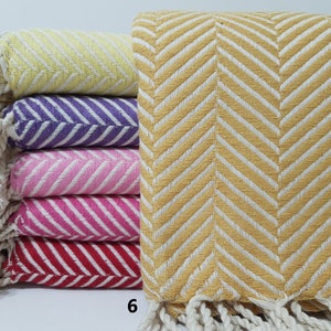 May include: A stack of six cotton towels with a herringbone pattern. The towels are in various colors, including yellow, purple, pink, and red. The towels have white fringe on the ends.