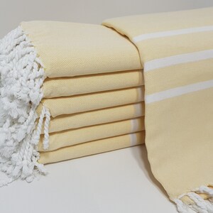 May include: A stack of yellow Turkish towels with white stripes and white fringe. The towels are folded and stacked on a white surface.