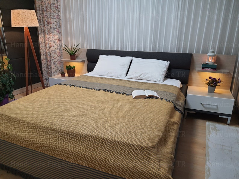 May include: A bedroom with a bed with a yellow and brown patterned blanket, two white pillows, and a book on top of the blanket. There are two nightstands with lamps on each side of the bed.