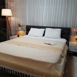 K&ouml;nnte beinhalten: Eine beige-wei&szlig;e gestreifte Wolldecke mit Fransen auf einem Bett mit wei&szlig;en Kissen und einem Buch auf der Decke. Das Bett steht in einem Schlafzimmer mit einem Nachttisch und einer Lampe.