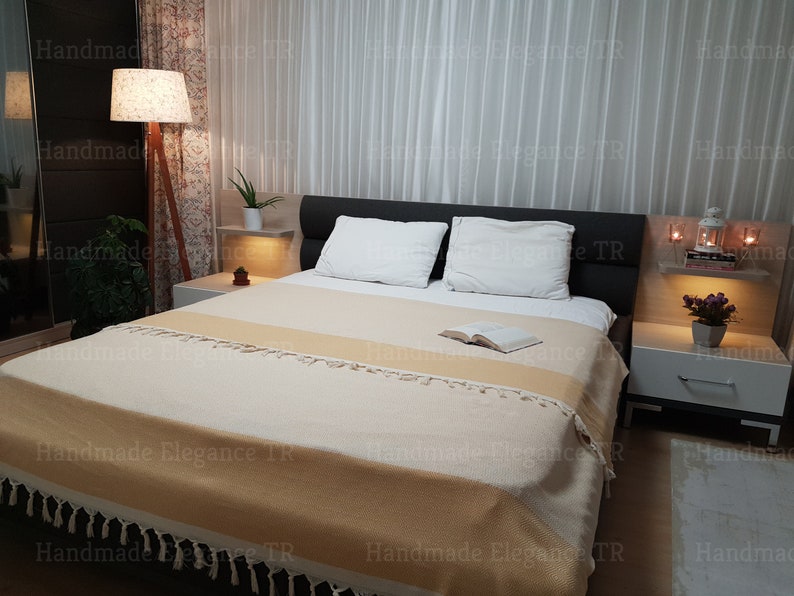 K&ouml;nnte beinhalten: Eine beige-wei&szlig;e gestreifte Wolldecke mit Fransen auf einem Bett mit wei&szlig;en Kissen und einem Buch auf der Decke. Das Bett steht in einem Schlafzimmer mit einem Nachttisch und einer Lampe.