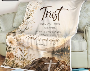 May include: A cream-colored throw blanket with a scenic landscape design and the word "Trust" in a brown script. The blanket features a lake, mountains, and a cross, with the text "God is our refuge" and Psalm 62:8.