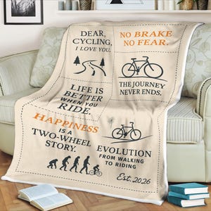 May include: A cream-colored throw blanket with black and orange text and illustrations related to cycling. The blanket features phrases like "Dear Cycling, I love you," and "Happiness is a two-wheel story." The blanket also has bicycle graphics.