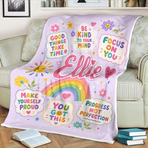 May include: A light purple throw blanket featuring the name Ellie in large pink script, surrounded by colorful illustrations of rainbows, flowers, suns, and hearts, along with text bubbles reading Good Things Take Time, Be Kind To Your Mind, Focus On You, Make Yourself Proud, You Got This, and Progress Not Perfection.