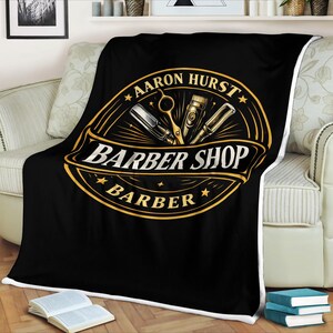 Może przedstawiać: Czarny koc z białym obramowaniem z logo zakładu fryzjerskiego w kolorze złotym i czarnym. Logo zawiera tekst "Aaron Hurst", "Barber Shop" i "Barber", a także ilustracje narzędzi fryzjerskich.