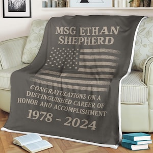 Puede incluir: Una manta de franela gris con un borde blanco. La manta presenta un diseño de bandera estadounidense desgastado con el texto "MSG ETHAN SHEPHERD" y "CONGRATULATIONS ON A DISTINGUISHED CAREER OF HONOR AND ACCOMPLISHMENT 1978 - 2024".