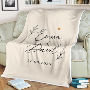 Könnte beinhalten: Eine cremefarbene Fleece-Decke mit einer schwarzen, kursiv geschriebenen Aufschrift "Emma & David" und dem Datum "22.08.2025". Ein kleines goldenes Herz befindet sich über den Namen.