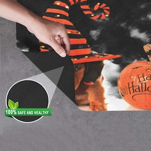 Pode incluir: Um tapete de entrada com tema de Halloween com um dachshund usando um chap&eacute;u de bruxa e uma ab&oacute;bora com o texto "Happy Halloween". O tapete tem um fundo preto com nuvens brancas e uma borda preta.