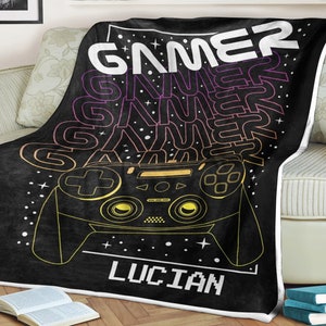Op de afbeelding: Een zwarte fleece deken met een gamer-thema. De deken heeft een gestileerde afbeelding van een videogamecontroller met het woord "GAMER" dat in een kleurverloop wordt herhaald. De naam "LUCIAN" is in een pixellettertype onderaan de deken gedrukt.