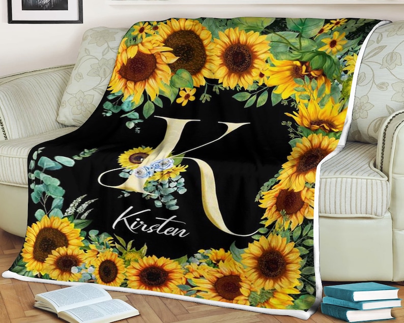 May include: Black fleece blanket with a white letter K and the name Kirsten printed on it. The blanket is decorated with a border of sunflowers and green leaves.