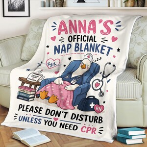 May include: A white throw blanket featuring a cartoon goose dressed as a nurse, sitting in a blue armchair. The text reads: ANNA'S OFFICIAL NAP BLANKET, PLEASE DON'T DISTURB UNLESS YOU NEED CPR. Includes a stethoscope, books, and a medical bag.