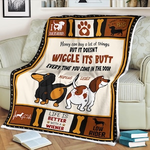 May include: A cream-coloured blanket with dachshund illustrations and the text "Money can buy a lot of things BUT IT DOESN'T WIGGLE ITS BUTT EVERY TIME YOU COME IN THE DOOR." The blanket has a black and brown border and includes the words "I Dachshunds", "Life is better with a wiener", and "Low Rider".