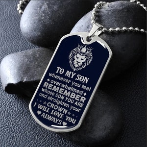 May include: A silver dog tag necklace with a blue background and silver text. The text reads "To My Son whenever you feel overwhelmed Remember whose son you are and straighten your crown I will love you always". A silver lion with a crown is in the top left corner of the tag.