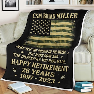 Puede incluir: Manta negra con un diseño de bandera estadounidense en camuflaje. La manta tiene texto que dice "CSM BRIAN MILLER" y "HAPPY RETIREMENT 26 YEARS 1997-2023". La manta está sobre un sofá.