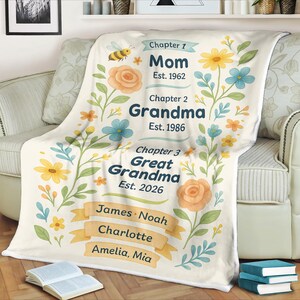 May include: A cream-coloured blanket with floral and bee illustrations. The blanket features text that reads "Mom Est. 1962", "Grandma Est. 1986", and "Great Grandma Est. 2026". Names are listed below the text. The blanket is draped over a sofa.