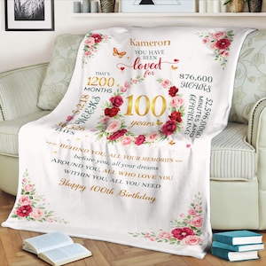 Pode incluir: Um cobertor branco com um design floral e texto. O cobertor tem uma coroa de flores em torno do número 100, com as palavras "Kameron, You Have Been Loved For" acima. O cobertor também inclui o texto "Happy 100th Birthday".