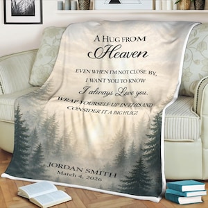 May include: A soft, white blanket with a forest scene and the text "A HUG FROM Heaven." The blanket includes the message "I always Love you" and a name and date. The blanket is draped over a sofa.