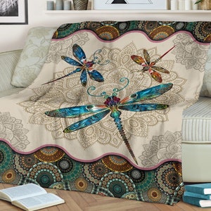 May include: A cream-colored throw blanket with a mandala pattern and three colorful dragonfly designs. The blanket has a pink and brown border with a repeating pattern of circles.