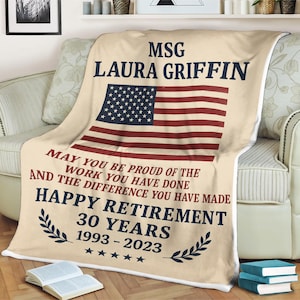 Puede incluir: Una manta beige con un diseño de la bandera estadounidense. La manta tiene texto que dice "MSG LAURA GRIFFIN" y "HAPPY RETIREMENT 30 YEARS 1993-2023". La manta está sobre un sofá.