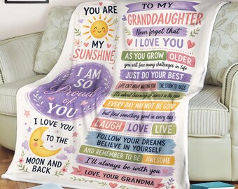 May include: A white fleece blanket featuring a colorful printed message to a granddaughter. The text includes phrases like You Are My Sunshine, I Love You To The Moon And Back, and Love, Your Grandma, accompanied by illustrations of a sun and moon.