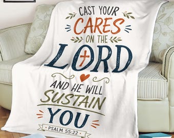 May include: A white throw blanket featuring the text CAST YOUR CARES ON THE LORD AND HE WILL SUSTAIN YOU PSALM 55:22 in dark blue, orange, and green lettering, with a small orange cross inside the letter O and a small orange heart below it.