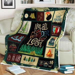 May include: A colourful fleece blanket with a camping theme. It features a variety of images, including trees, mountains, a compass, a tent, and a campfire. The text on the blanket reads "Not all who wander are lost", "Gone Hiking", "Born to Hike", "Mountains Calling", and "Take me to the trees".