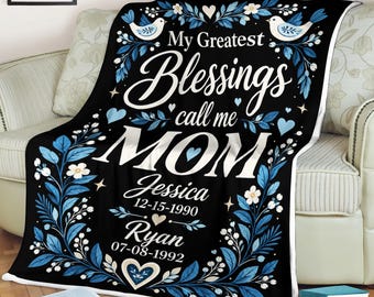 Pode incluir: Uma manta de lã preta com ramos florais azuis e brancos, dois pássaros e corações. O texto diz: My Greatest Blessings call me MOM, Jessica 12-15-1990 e Ryan 07-08-1992. O design inclui flores brancas e folhas azuis sobre um fundo preto.