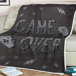 May include: A black and gray throw blanket with the words "Game Over" in a bold, textured font. The blanket features a pixelated design with game controller icons.