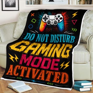 May include: A black fleece blanket featuring a white video game controller graphic at the top, surrounded by colorful geometric shapes and lightning bolts. The text reads DO NOT DISTURB GAMING MODE ACTIVATED in large, bold, distressed yellow, pink, and orange letters.