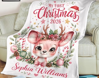 May include: A white fleece blanket featuring a central illustration of a fawn with antlers, decorated with holly, pine branches, candy canes, and ornaments. The text reads My First Christmas 2026 and the name Sophia Williams in red script.