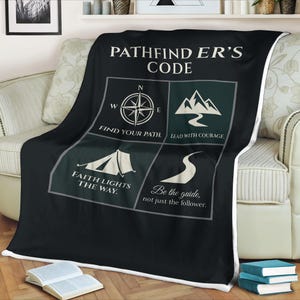 Puede incluir: Manta negra con texto blanco "PATHFINDER'S CODE". La manta presenta una brújula, montañas, una tienda de campaña y un camino sinuoso. También se incluyen las frases "Find Your Path", "Lead with Courage", "Faith Lights the Way" y "Be the guide, not just the follower".