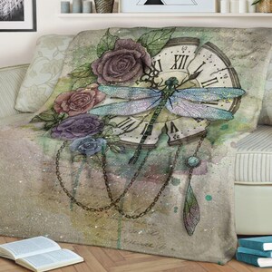 May include: A vintage-style throw blanket with a watercolor design featuring a dragonfly, roses, and a clock face. The background is a soft green with hints of pink and blue.