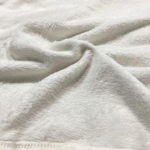 May include: Close-up of a white, soft, plush fabric. The fabric has a fuzzy texture and is folded in a way that shows its softness.