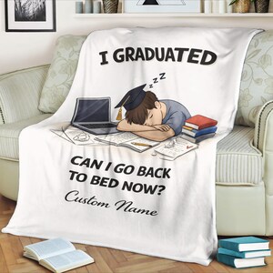 以下が含まれることがあります： 「I GRADUATED」と「CAN I GO BACK TO BED NOW？」の文字が入った白いフリースブランケット。本とノートパソコンの上で寝ている人の漫画イラストが描かれています。ブランケットはソファに掛けられています。