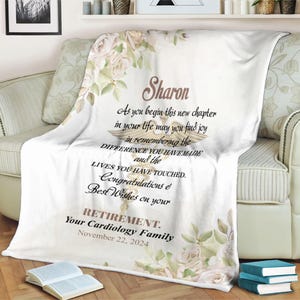 Puede incluir: Una manta de forro polar blanca con un símbolo médico dorado y un diseño floral. El texto dice "Sharon As you begin this new chapter in your life may you find joy in remembering the DIFFERENCE YOU HAVE MADE and the LIVES YOU HAVE TOUCHED. Congratulations & Best Wishes on your RETIREMENT. Your Cardiology Family November 22, 2024".