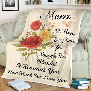 May include: Cream-colored blanket with a floral design featuring red and yellow roses, green leaves, and butterflies. The blanket has the words "Mom," "We Hope Every Time You Snuggle This Blanket It Reminds You How Much We Love You."